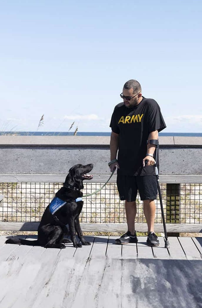 A man wearing an 'ARMY' T-shirt and using a cane stands on a boardwalk overlooking the ocean with a black service dog sitting next to him, wearing a blue service dog vest.
