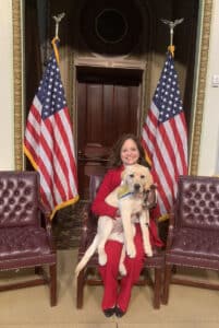 A woman in a red suit sits between two American flags, holding a yellow Labrador Retriever wearing a yellow and blue Canine Companions vest.