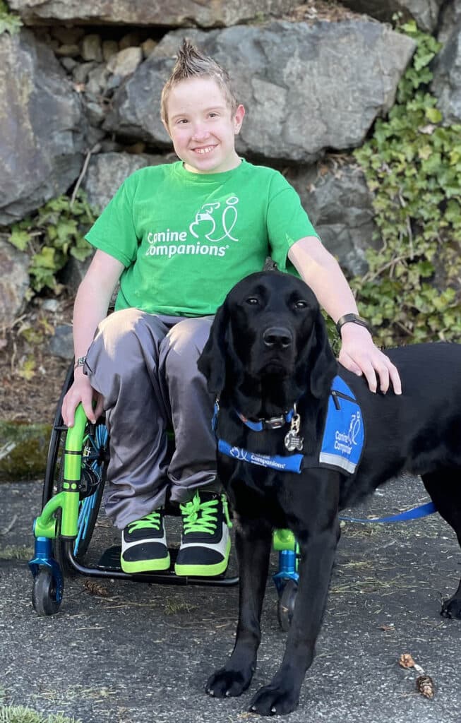 A person in a green Canine Companions t-shirt sitting in a wheelchair, smiling and posing with a black lab in a Canine Companions vest.