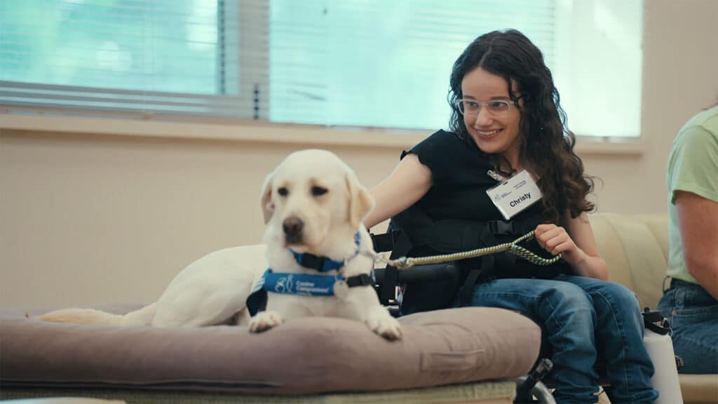 A young woman in a wheelchair, wearing glasses and a name tag, smiles while petting a service dog lying on a cushion.