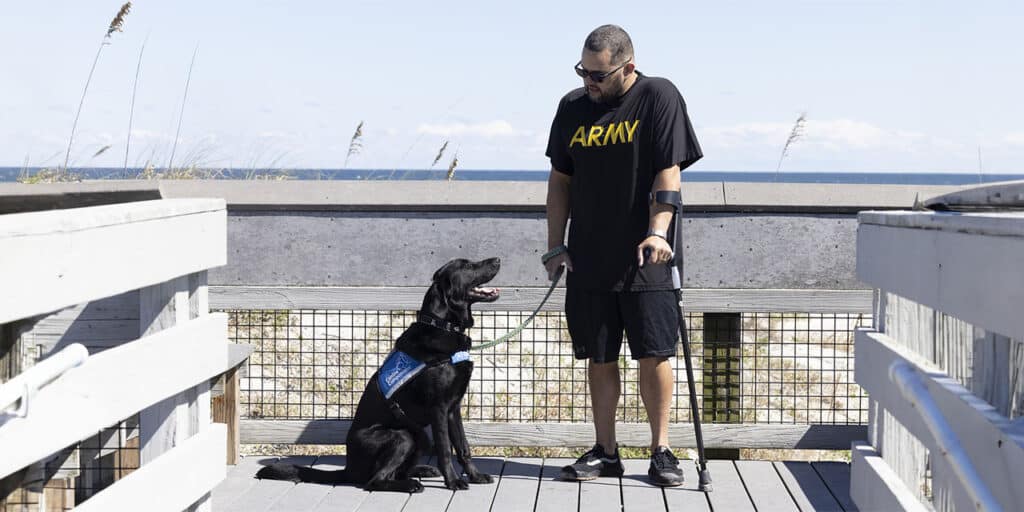 A man wearing an 'ARMY' T-shirt stands with his black service dog on a boardwalk by the beach.