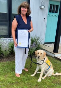 A woman holding an official statement stands next to a seated yellow Labrador wearing a "Canine Companions" vest. They are outside a house with a light blue door.