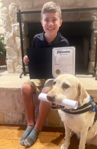 A boy sitting by a fireplace, holding an open folder with a proclamation inside, while a Labrador Retriever in a service dog vest sits beside him holding a rolled paper in its mouth.