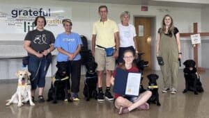 A group of seven people, some with guide dogs, posing indoors. A young girl in front is holding a certificate. The background has a wall with words like "grateful" and "appreciate" displayed.