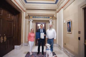 Three people and a service dog posing in front of a doorway labeled "Governor"; one of the people is holding a certificate.