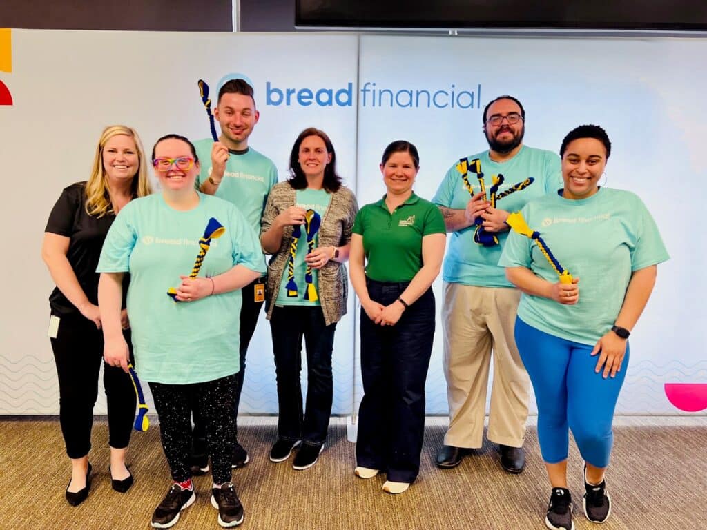 A group of seven people standing in front of a "bread financial" backdrop holding colorful braided ropes. Some are wearing light blue "bread financial" T-shirts, and one is wearing a green shirt. They are all smiling at the camera.