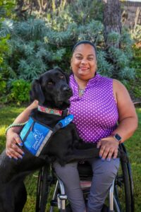 Woman in a wheelchair with a black assistance dog wearing a "Canine Companions" vest, posing outdoors.