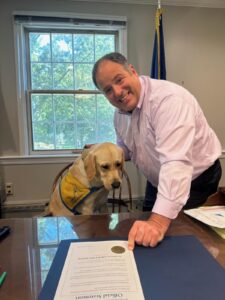 A man pointing towards a document on a desk with a yellow Labrador retriever wearing a service dog vest beside him.