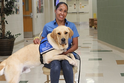 A woman seated on a chair, smiling, with a yellow Labrador wearing a blue "Canine Companions" vest in her lap.