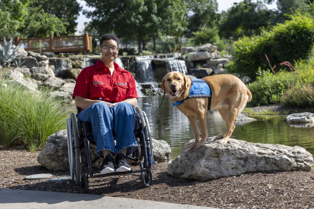 A person in a wheelchair poses with a service dog near a pond with a waterfall in the background.