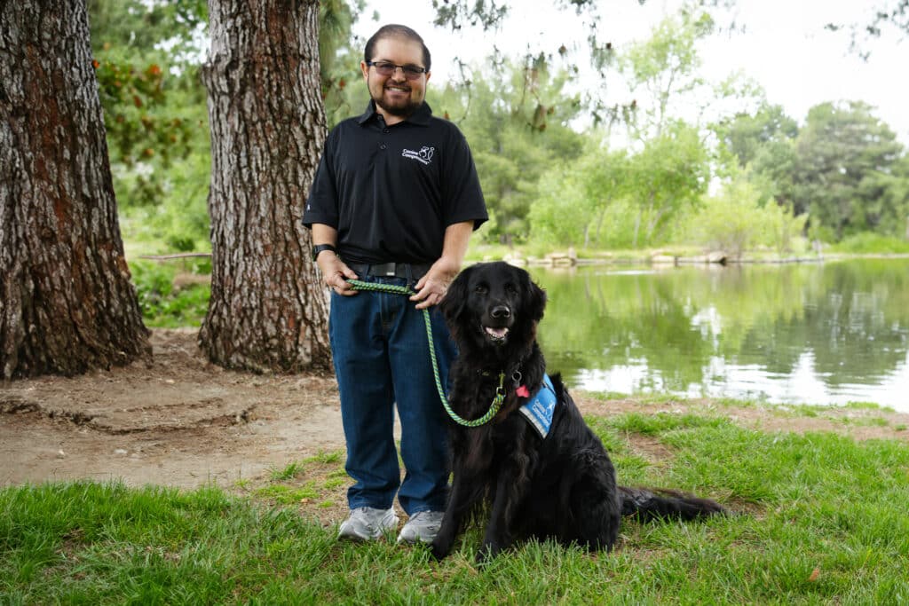 A man standing outdoors near a pond, holding the leash of a black service dog wearing a blue vest, with trees in the background.