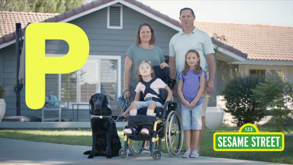 A family consisting of two adults, two children, a black service dog, and a wheelchair user posing in front of a house with a large, yellow "P" and the Sesame Street logo.