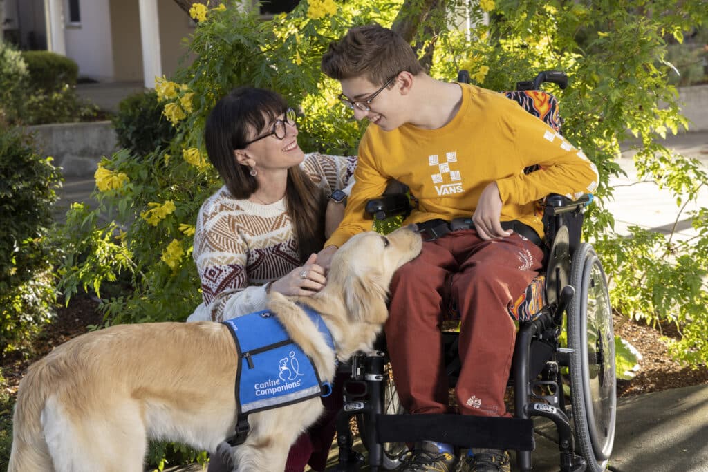 A woman crouches next to a young person in a wheelchair, both smiling at each other, while a service dog wearing a "Canine Companions" vest rests its head on the young person's lap. They are outside with green foliage and yellow flowers in the background.