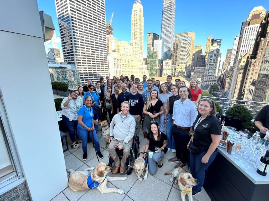 Group of smiling people posing on a rooftop with several guide dogs, overlooking a city skyline.