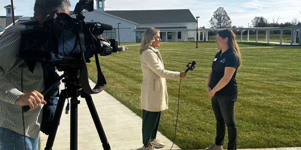 A TV reporter with a microphone interviews a woman outside on a grass lawn, while a cameraman films the scene.