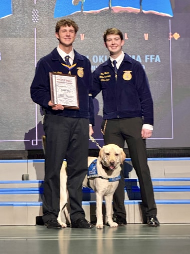 Two young men in blue jackets standing on stage next to a yellow service dog. One is holding a framed certificate.