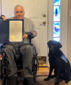 A man in a wheelchair holding a certificate sits next to a black service dog on the floor, with a door in the background.