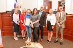 A group of people posing in a formal room with wood paneling and a flag, holding a certificate, with a service dog lying on the floor in front.