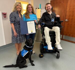 Three people posing indoors with a service dog wearing a blue vest. One woman is holding a proclamation document, another is standing beside her, and a man is in a wheelchair.