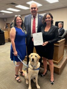 Two women and a man dressed in formal wear pose together in an office setting, with the man holding a certificate. A service dog sits in front of them.