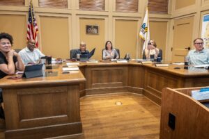 A group of six people sitting at a city council meeting table with nameplates, the American flag, and a city flag behind them.