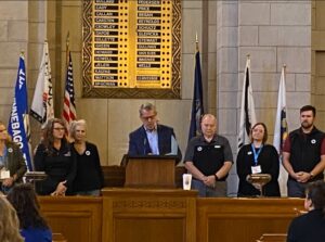 A group of people standing behind a podium in a formal room with flags and a board listing names in the background.