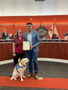 Two people and a service dog in front of a city council meeting with an official proclamation.