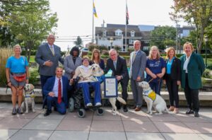 A group of eleven people, some holding service dogs, stands in an outdoor setting. One person in a wheelchair holds a dog in their lap. The group poses with a framed proclamation. Trees and flags are visible in the background.