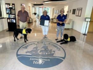 Three people holding service dogs standing around the James City County logo inside a building, with one person holding a certificate.