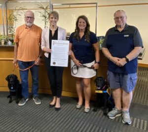 Group photo of four people standing in an office, two of them holding leashes with black service dogs. The woman in the center holds a Proclamation document recognizing National Service Dog Month.