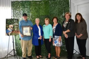 Group of six adults standing in front of a greenery backdrop, one holding a certificate, next to a poster of a dog in a service vest.