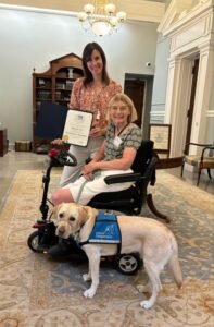 Two women, one sitting in a mobility scooter and one standing, both smiling and holding a certificate, with a service dog wearing a "Canine Companions" vest in the foreground.