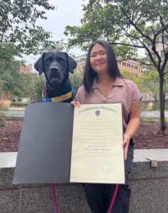 A woman and a black dog holding a proclamation document outside.