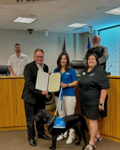 A group of people posing with a document in a meeting room; a woman is holding a black service dog with a "Canine Companions" vest.