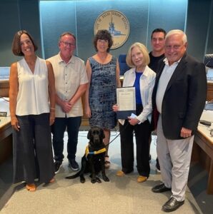 A group of five adults standing indoors, smiling at the camera. One person holds a certificate while a black Labrador service dog sits in front.