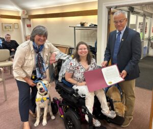 A woman in a beige jacket is standing beside a yellow Labrador wearing a blue service dog vest. Another woman in a wheelchair with a service dog next to her is receiving a certificate from a man in a dark suit. People are seated in the background of the room.