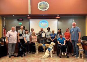 A group of people and service dogs posing together in a room with "Rohnert Park, California" displayed on the wall and screen in the background.