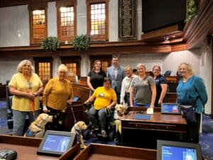 A group of people, some with service dogs, standing in a legislative chamber with wooden desks and a display of names on the wall.