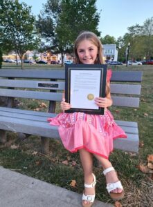 Young girl sitting on a bench outdoors, holding a framed certificate and smiling.