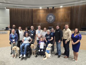 A diverse group of people, including individuals in wheelchairs and service dogs, posing in front of a "City of Sunrise Florida" emblem in a council chamber.