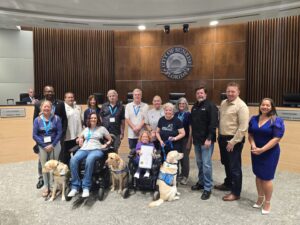 A group of people posing inside a city council chamber with a sign that reads "City of Sunrise Florida." Some individuals in wheelchairs are accompanied by service dogs. Behind them, officials are seated at a desk with nameplates.