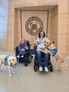 Two people in wheelchairs pose with service dogs in front of the "City of Sunrise, Florida" Commission Chambers.