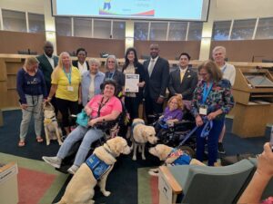 A group of people, some in wheelchairs and some with guide dogs, pose together indoors, holding a certificate.