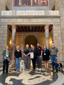 Group of seven people standing in a hallway with marble columns, accompanied by three service dogs.