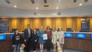 Group of seven people and two service dogs standing in front of a council chamber backdrop with "In God We Trust" sign and City of Yorba Linda logos.