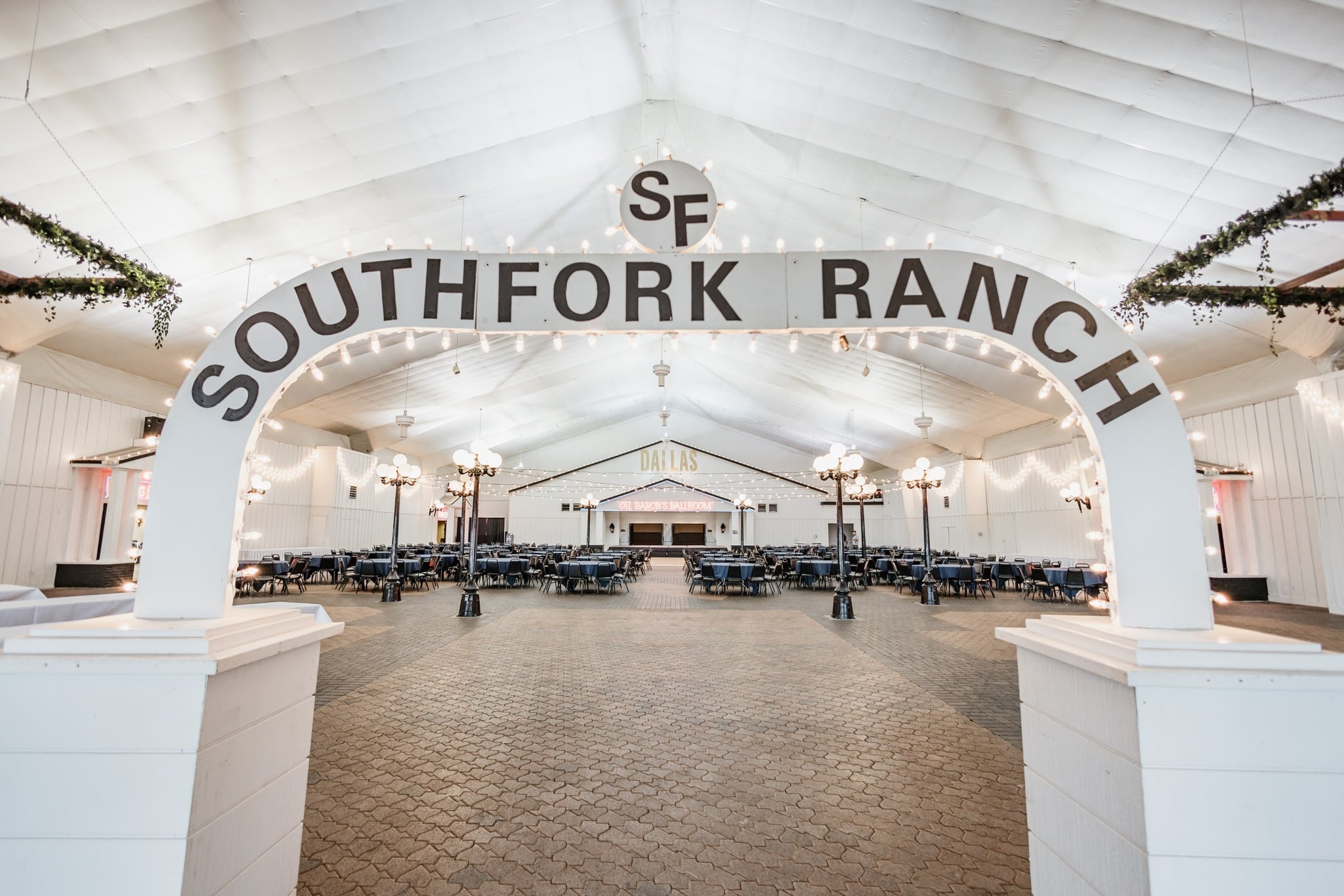 Archway sign reading "Southfork Ranch" leading to a large indoor event space with rows of tables and chairs, decorated with string lights and chandeliers.