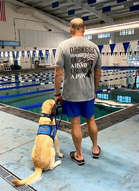 A man stands by an indoor pool with a service dog sitting beside him. The man's t-shirt reads, "In my darkest hour when I needed a hand, I found a paw."