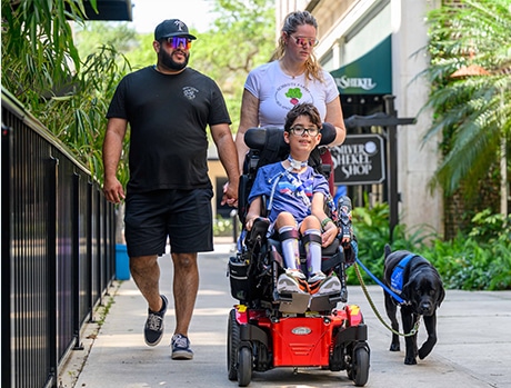 A family walking on a sidewalk; a child in a motorized wheelchair with a service dog alongside and two adults walking beside.