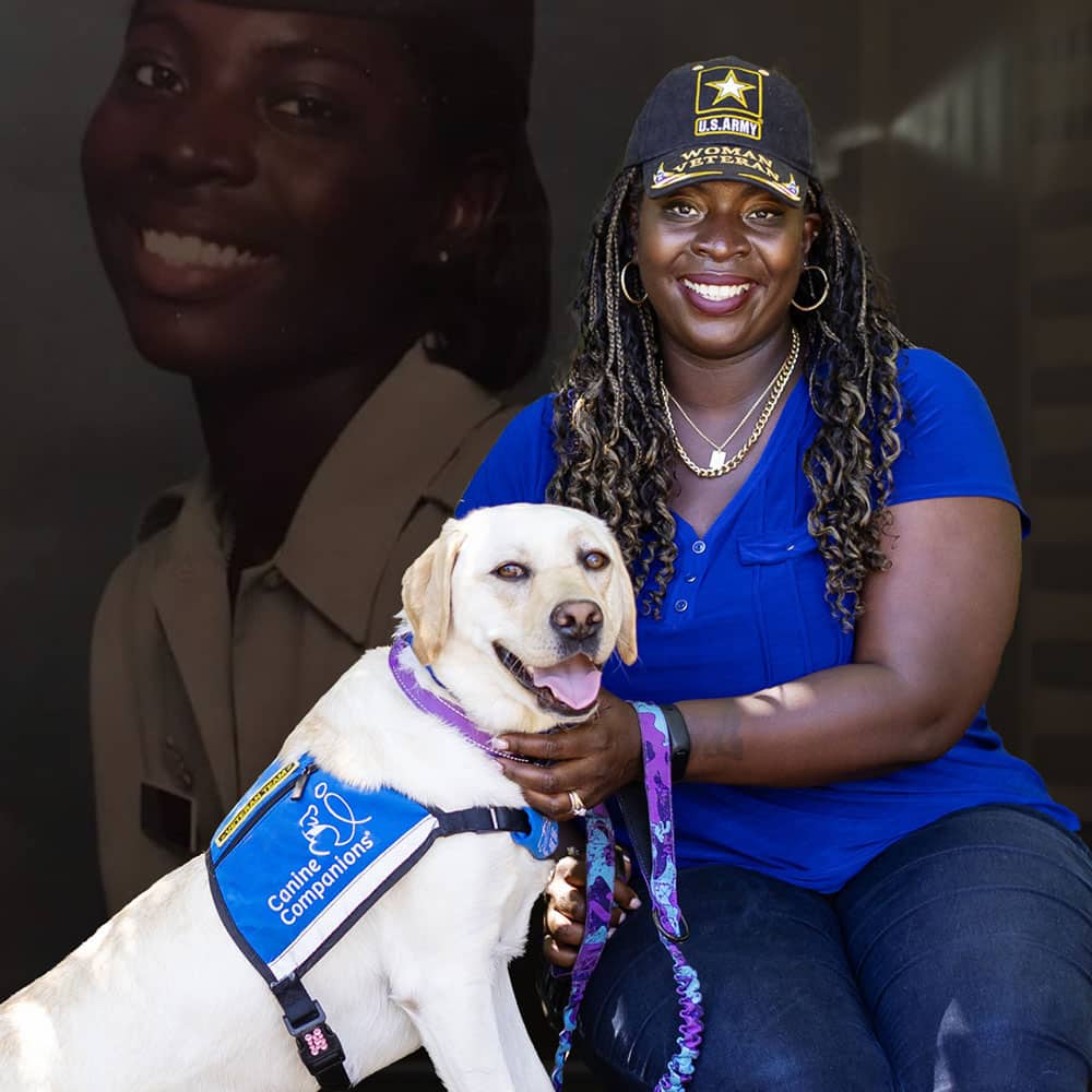 A woman wearing a U.S. Army veteran cap sitting with a service dog in a Canine Companions vest, smiling.