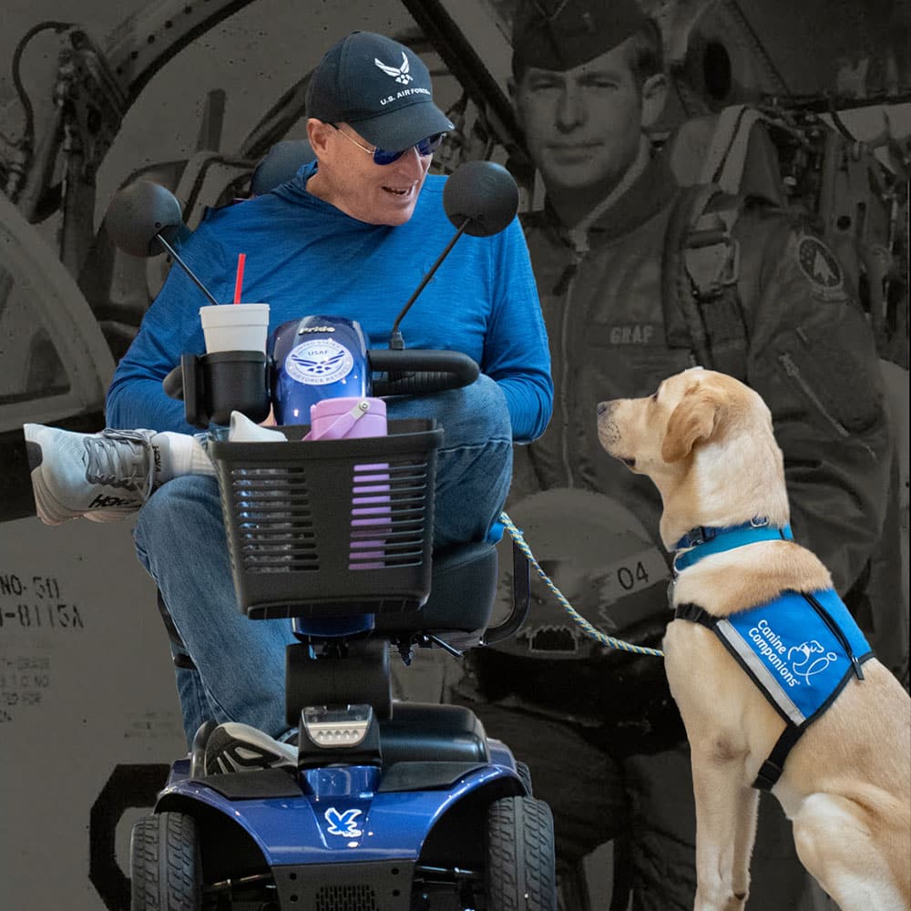 A man in a U.S. Air Force cap sits in a mobility scooter next to a service dog wearing a blue vest. A faded black and white photo of a pilot in uniform is in the background.